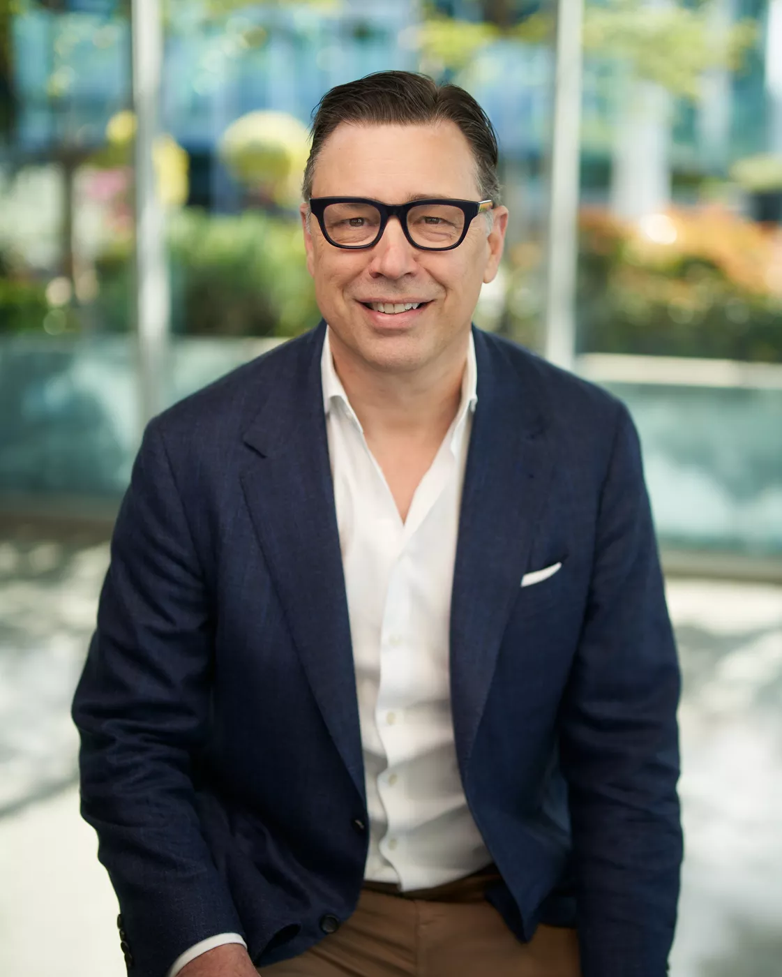 A professionally dressed man wearing a blue blazer and white shirt is seated outdoors. The setting features a modern glass building and vibrant greenery in the background. The scene conveys a professional and approachable mood with natural lighting.